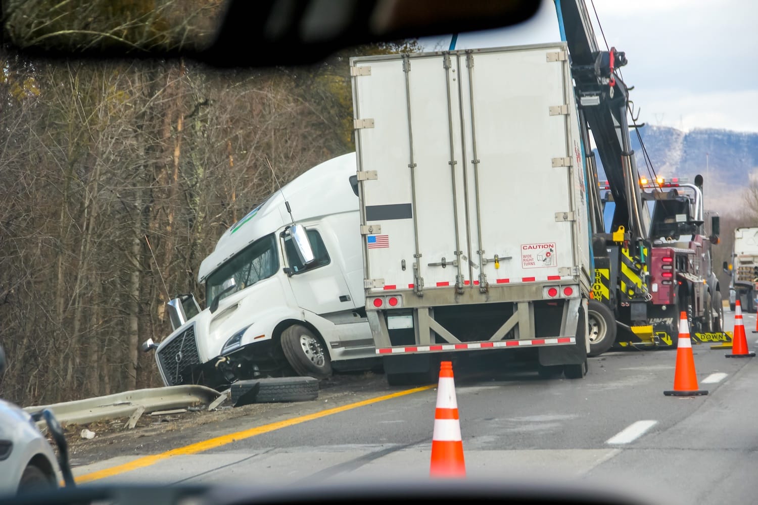 A jackknifed semi-truck on the side of a highway. A tow truck is pulling the truck back onto the road.