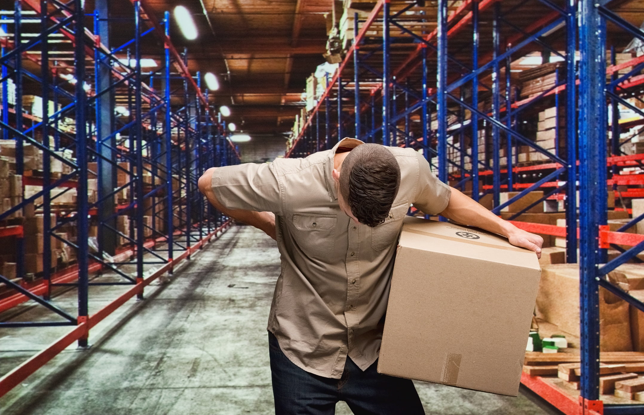 A man working in a warehouse holding his back in pain while carrying boxes.