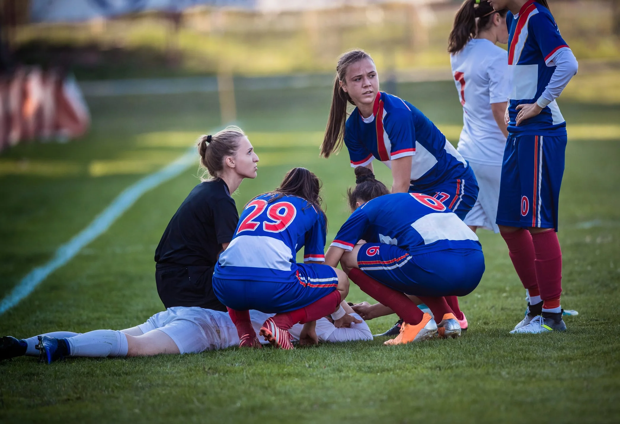 Female high school soccer players gather around an injured opponent who lies face-down on the field.