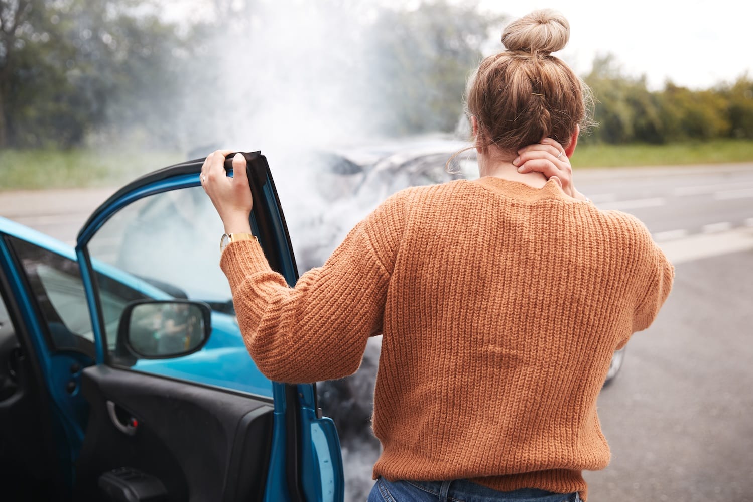Woman holding her neck next to a car after a car accident. Another car can be seen further ahead.